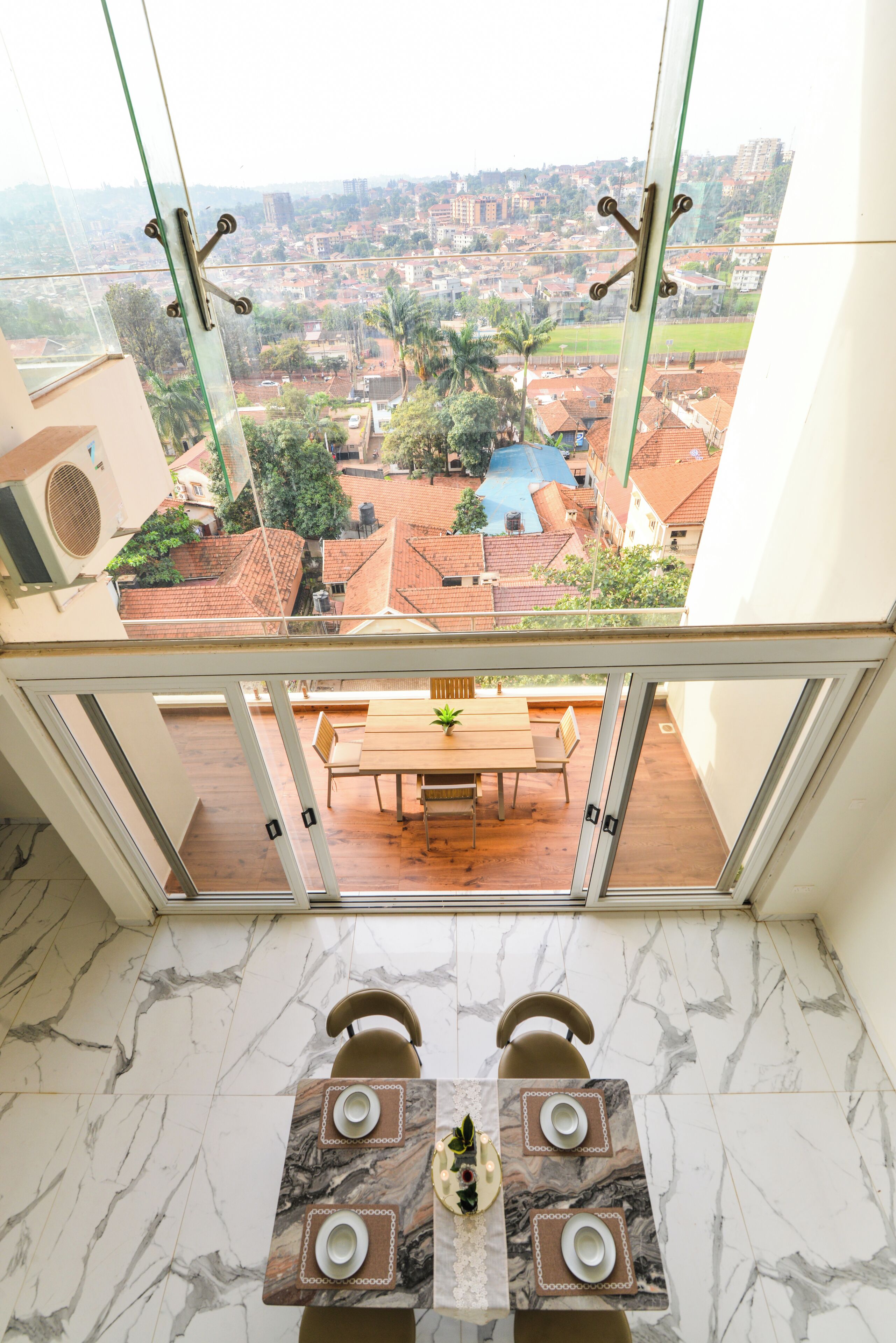 A high-rise apartment with a glass balcony overlooking a residential area, a dining table with chairs, and a view of a swimming pool.