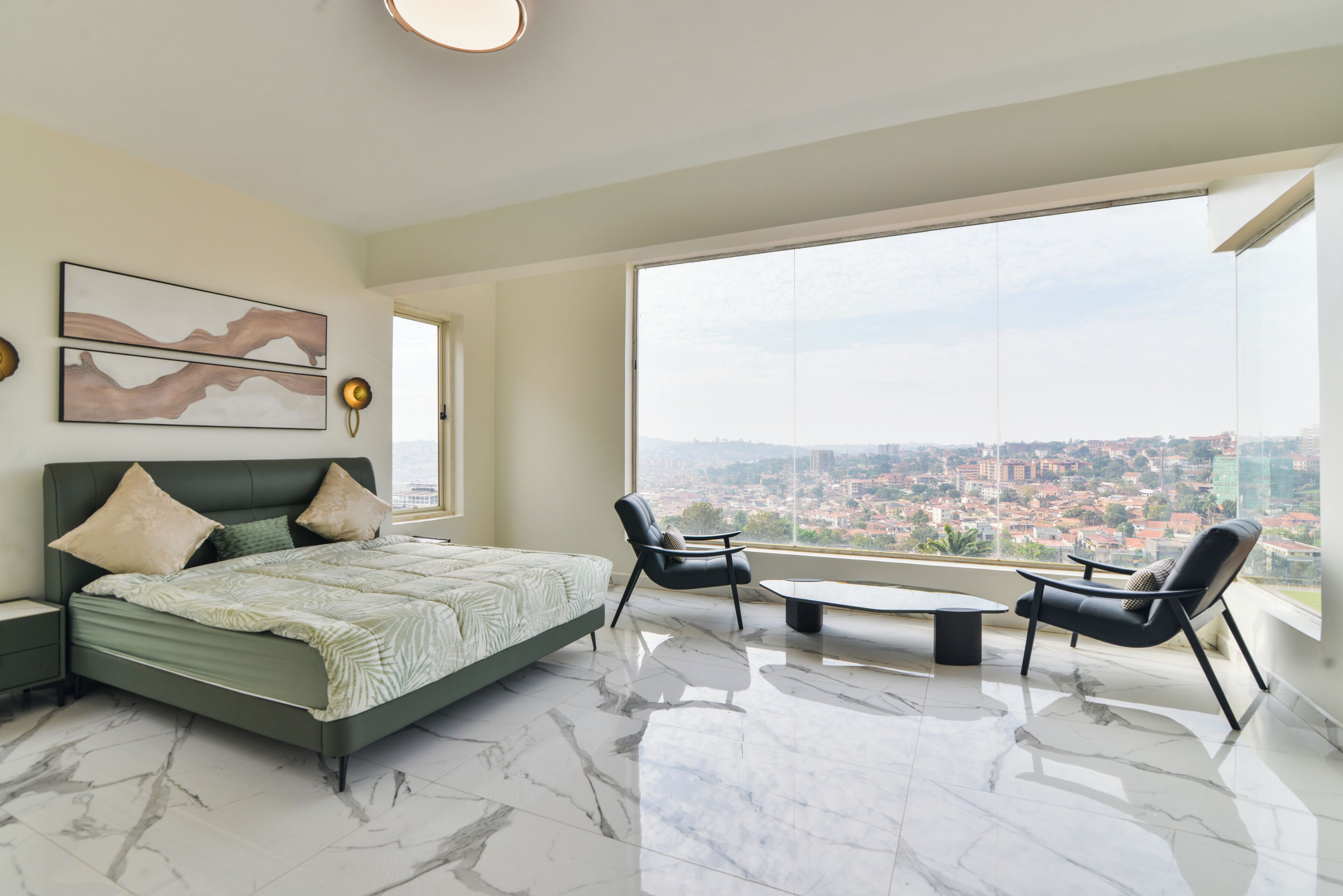 Un dormitorio moderno con una cama grande, dos sillas, una mesita y vista a la ciudad a través de un ventanal amplio.