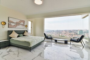 A modern bedroom with a large bed, a nightstand, a chair, a small table, and a city view through a large window. - Naperville Apartments Duplex (Kampala)