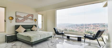A modern bedroom with a large bed, two chairs, a small table, and a city view.