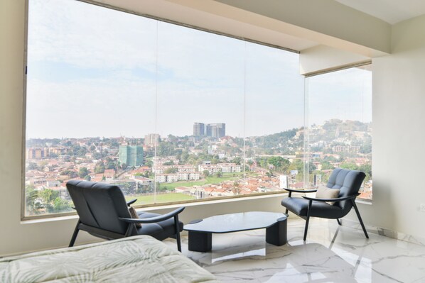 A modern room with a large window offering a city view, two black chairs, a small table, and a bed with a patterned bedspread.