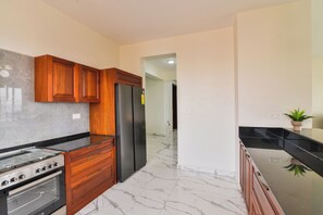 A kitchen with wooden cabinets, a stainless steel refrigerator, a black countertop, and a marble floor. - Naperville Apartments Duplex (Kampala)