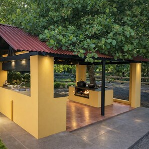 A covered outdoor cooking area with a fire feature, a sink, and a view of a garden.