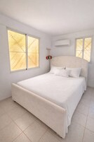 A bedroom with a large bed, two windows with blinds, an air conditioning unit, and a red clock on the wall.