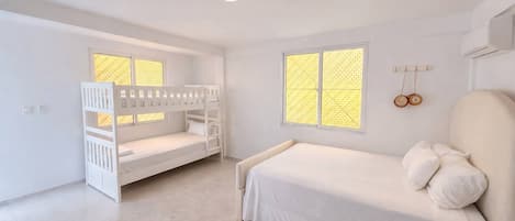 A bedroom with a bunk bed, a single bed, two windows with patterned glass, and a wall-mounted air conditioning unit.