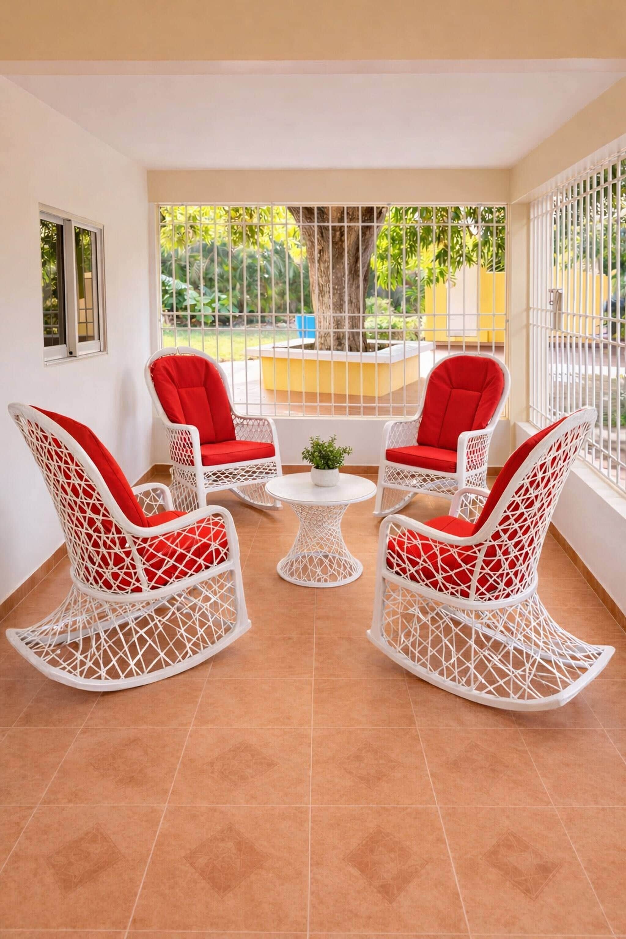 A room with white wicker rocking chairs and a small white table, large windows with a view of greenery and a yellow structure outside.
