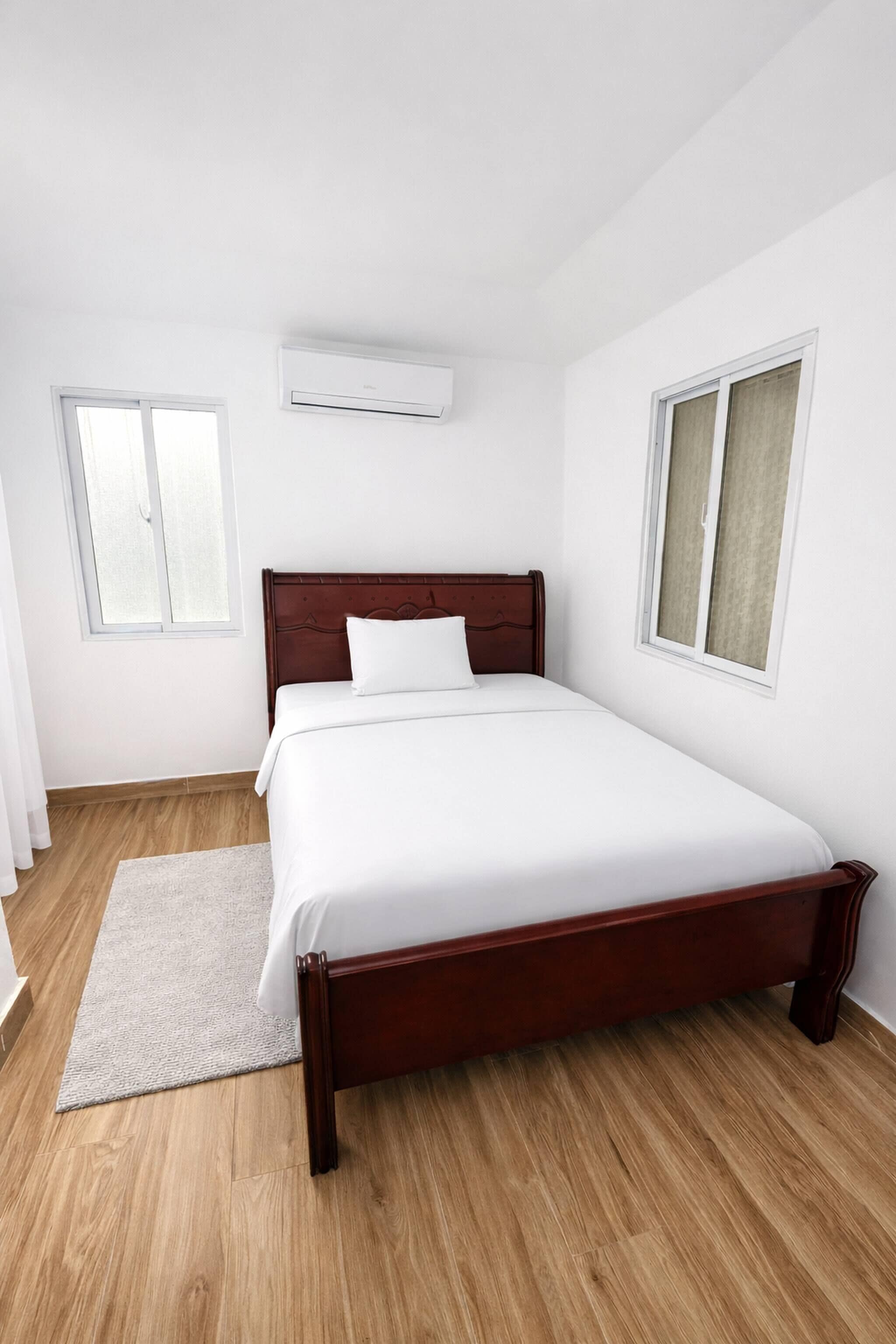 Un dormitorio con cama de madera, ropa de cama blanca, una alfombra blanca y dos ventanas con cortinas.