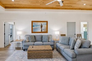 Una sala de estar moderna con un sofá gris, un reposapiés de mimbre y un cuadro de una persona en la playa.