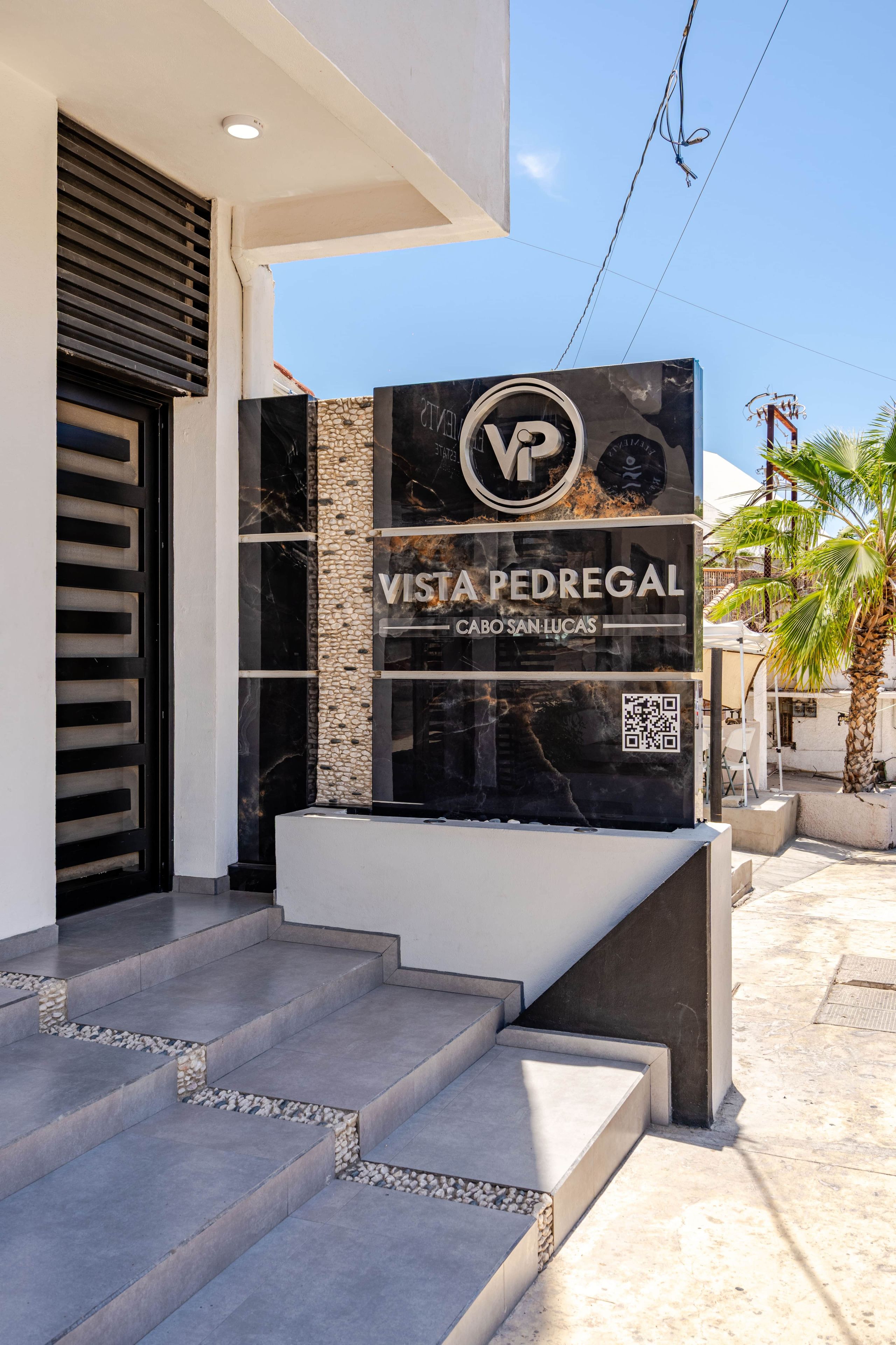 A modern building entrance with a sign for 'Vista Pedregal Cabo San Lucas' and a QR code.