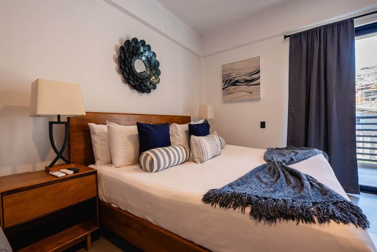 A hotel room with a bed, a nightstand, a lamp, a decorative wall piece, and a window with curtains.
