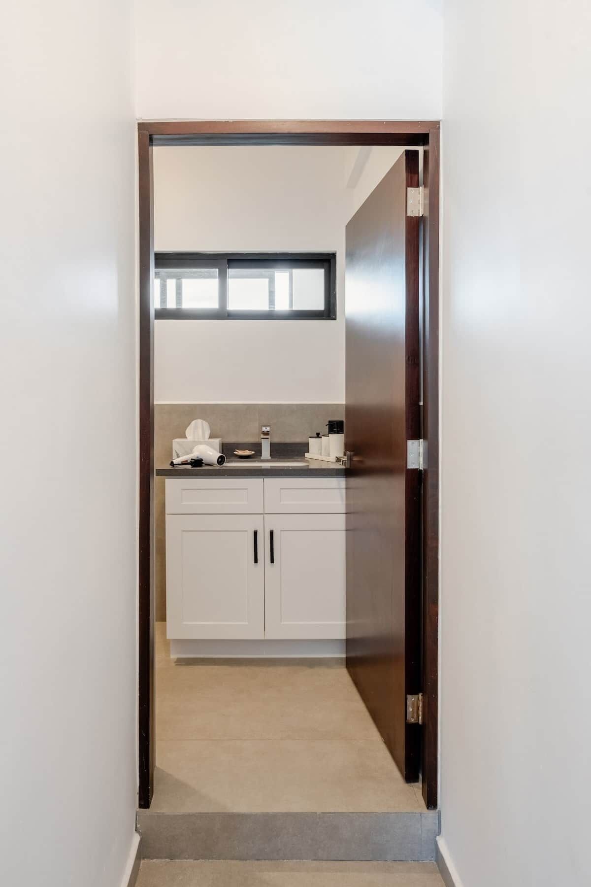 A narrow hallway leading to a kitchen with white cabinets, a countertop, and a window above the sink.