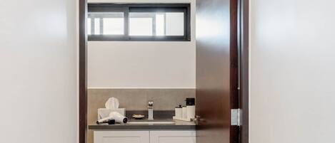 A narrow hallway leading to a kitchen with white cabinets, a countertop, and a window above the sink.