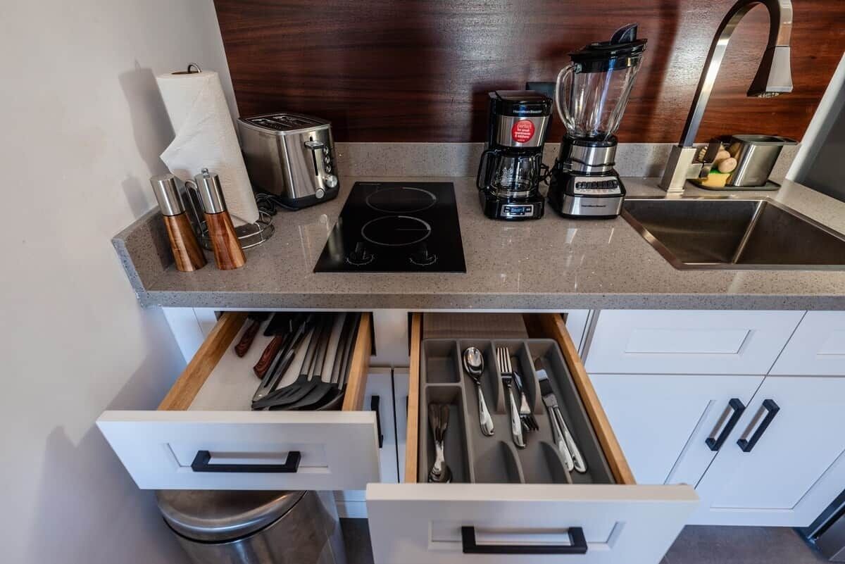 A kitchen with a toaster, coffee maker, blender, and a sink. There are utensils in a drawer and a paper towel roll.