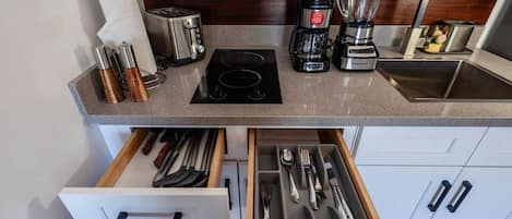 A kitchen with a toaster, coffee maker, blender, and a sink. There are utensils in a drawer and a paper towel roll.