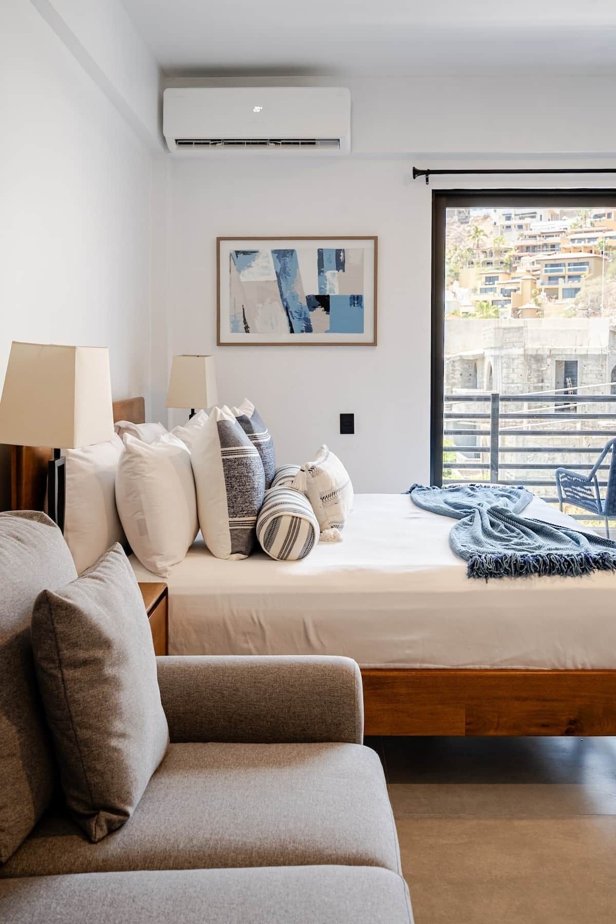 A modern bedroom with a bed, pillows, a sofa, a window with a view, and an air conditioning unit.