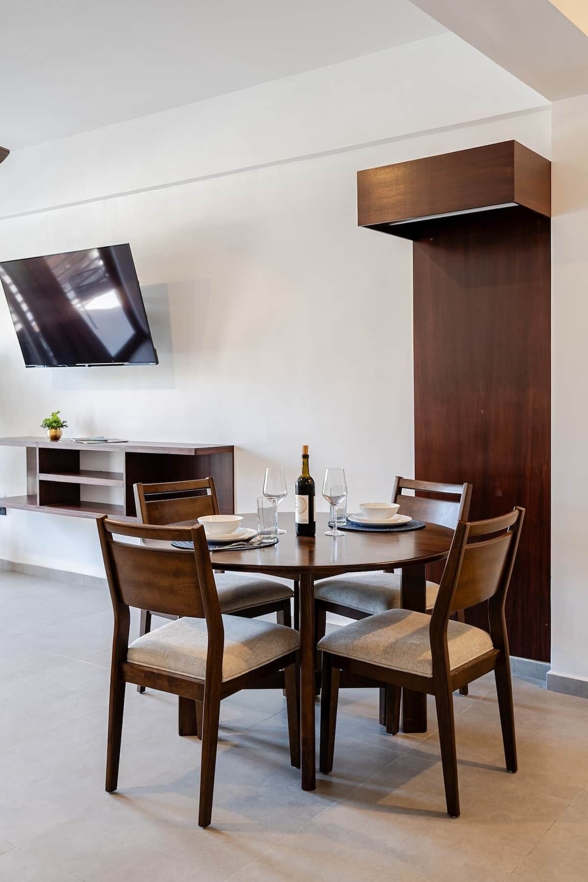 Un comedor moderno con una mesa redonda, cuatro sillas, un televisor montado en la pared y un mueble de madera.