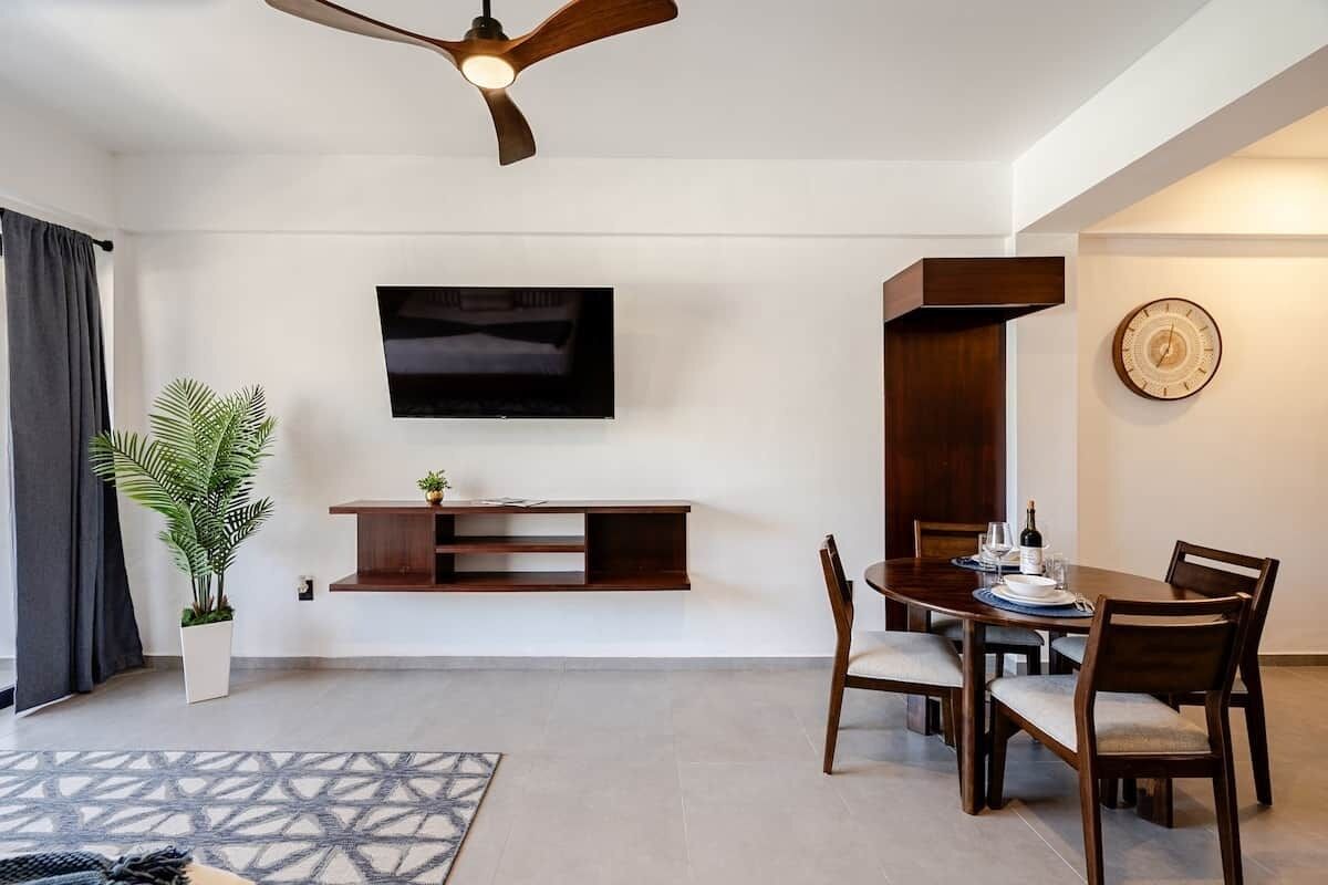 Un salon moderne avec un téléviseur à écran plat fixé au mur, une table à manger avec des chaises, un ventilateur de plafond et une plante en pot.