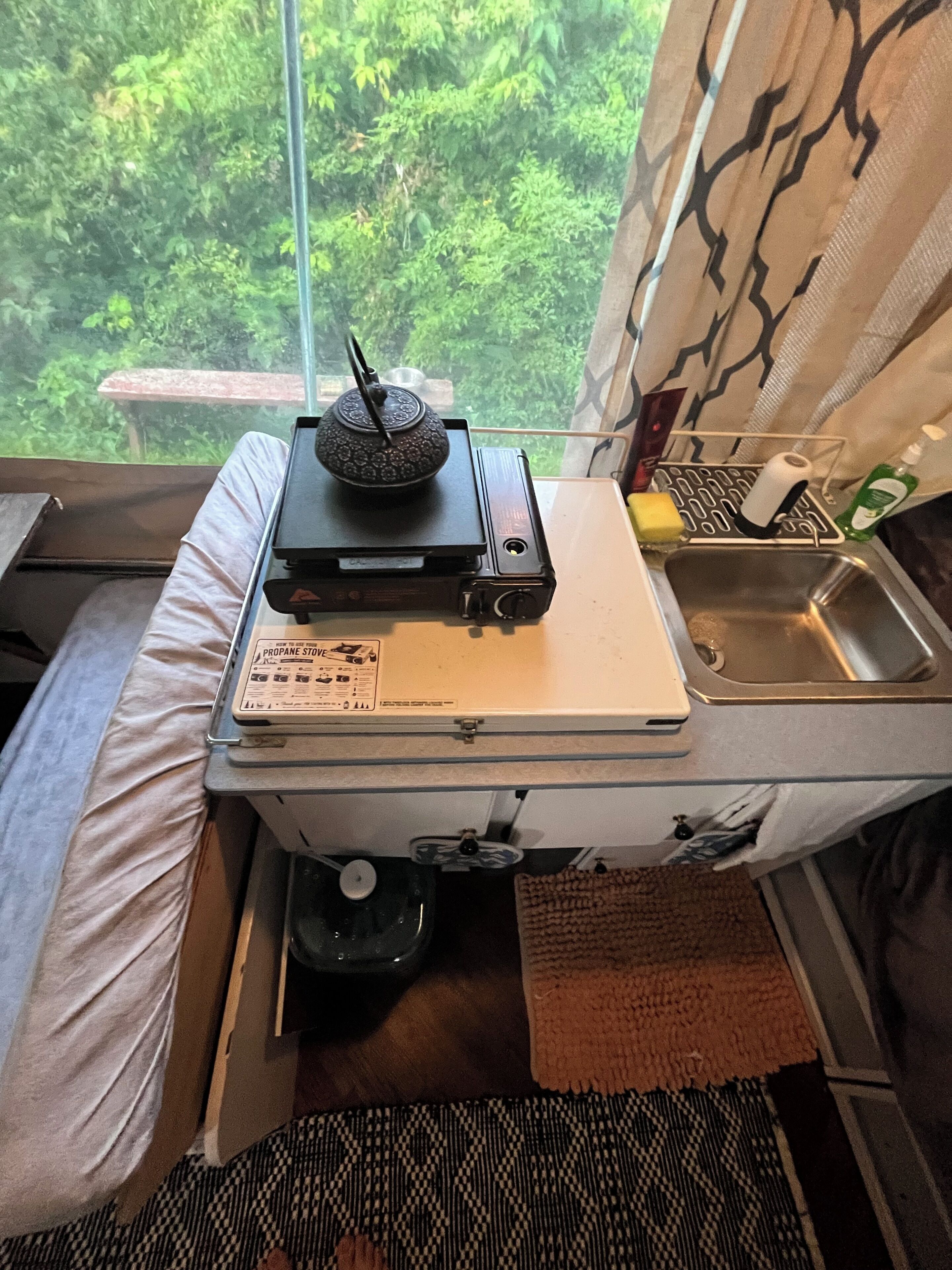 Een kleine keuken met kookplaat, spoelbak en een raam met uitzicht op groen.