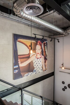 Ein modernes Interieur mit einem großen Kunstwerk, das ein Oldtimer-Auto zeigt, einer Treppe mit Glasgeländern und freiliegenden Leitungen an der Decke.