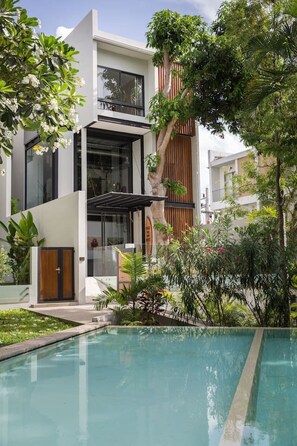 A modern building with a pool, surrounded by lush greenery and a clear sky.