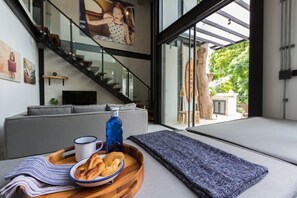 Un saló modern amb un sofà, un plat amb pastissos i una ampolla, i vista d'una escala i zona exterior.