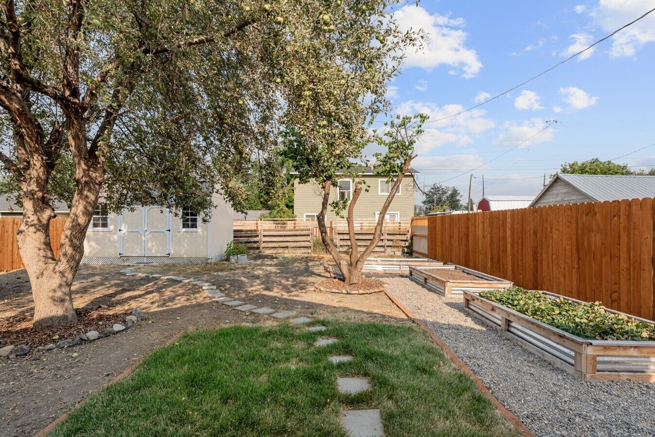 Un patio trasero cercado con un árbol, un edificio blanco y una valla de madera.