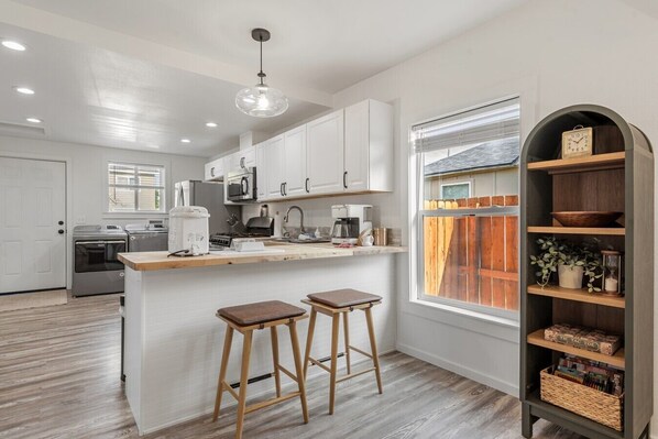 A modern kitchen with white cabinets, a wooden countertop, two stools, a coffee maker, and a window with blinds.