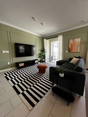 Un salon avec un téléviseur à écran plat, un canapé noir, un tapis à rayures et une plante en pot.