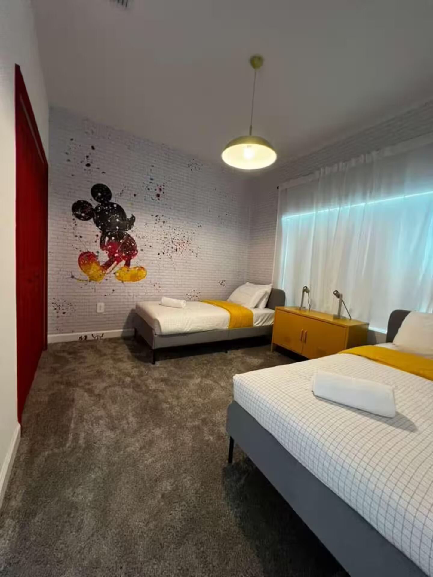A bedroom with two beds, a red wall, a white brick wall with a colorful mural, a yellow pendant light, and a window with curtains.