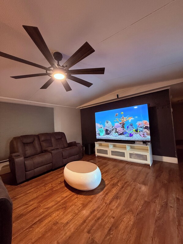 Un salon doté d’un ventilateur de plafond, d’un grand téléviseur à écran plat, d’un canapé en cuir marron et d’un pouf blanc.