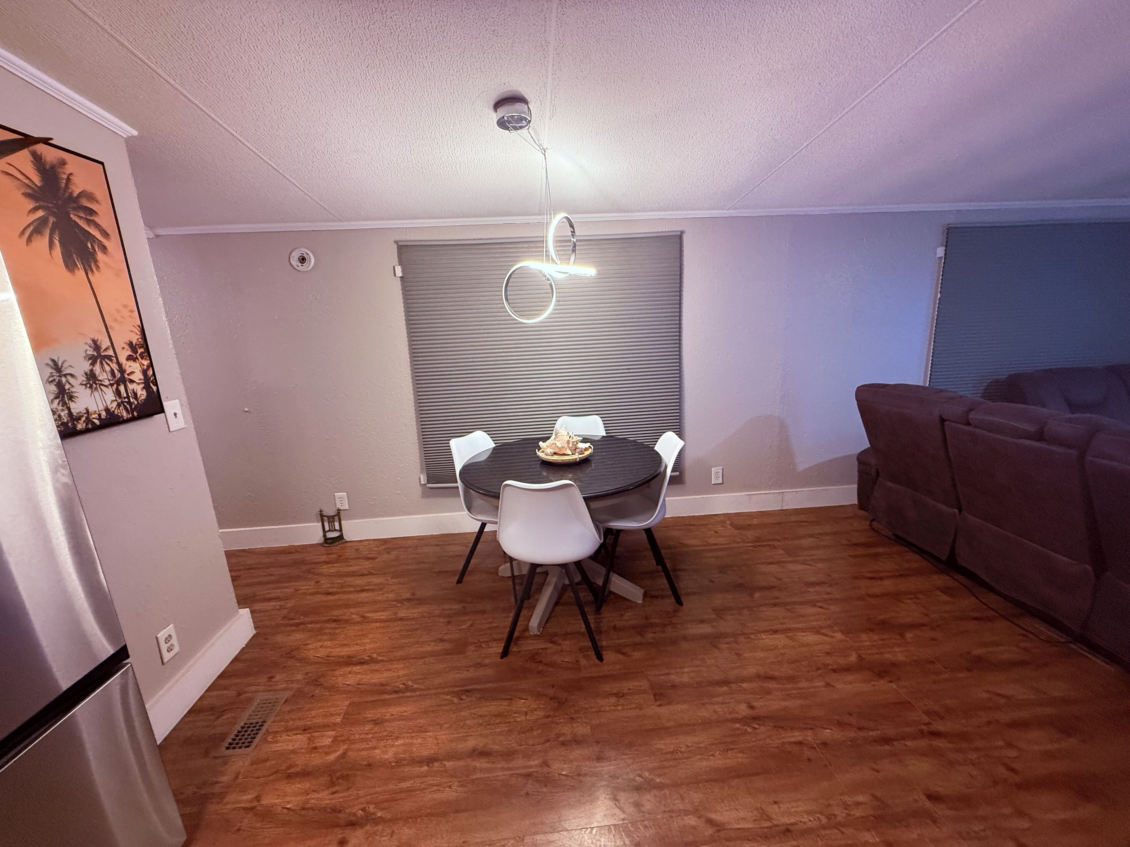 Un comedor con una mesa redonda y sillas, una lámpara de techo y un sofá marrón en una habitación con suelo de madera y techo blanco.