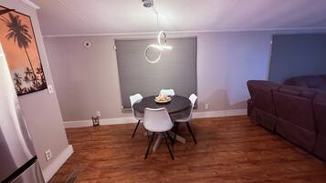 Un comedor con una mesa redonda y sillas, una lámpara de techo y un sofá marrón en una habitación con suelo de madera y techo blanco.