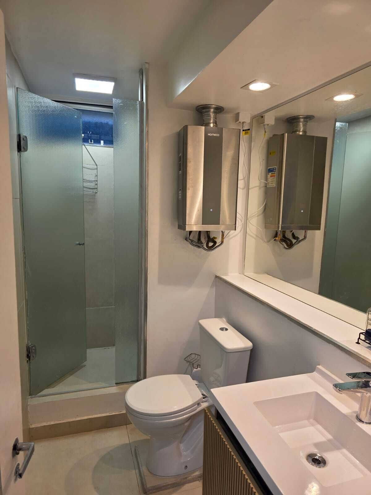 Uma casa de banho moderna com cabine de duche em vidro, sanita branca e lavatório com espelho em cima.