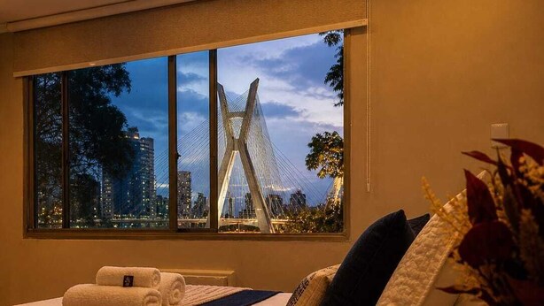 A hotel room with a large window, a bed with a white duvet, a bedside table with a lamp, and a view of a bridge and cityscape. - Duplex Apartment: Style and comfort just a few meters from Berrini & Shopping (São Paulo)