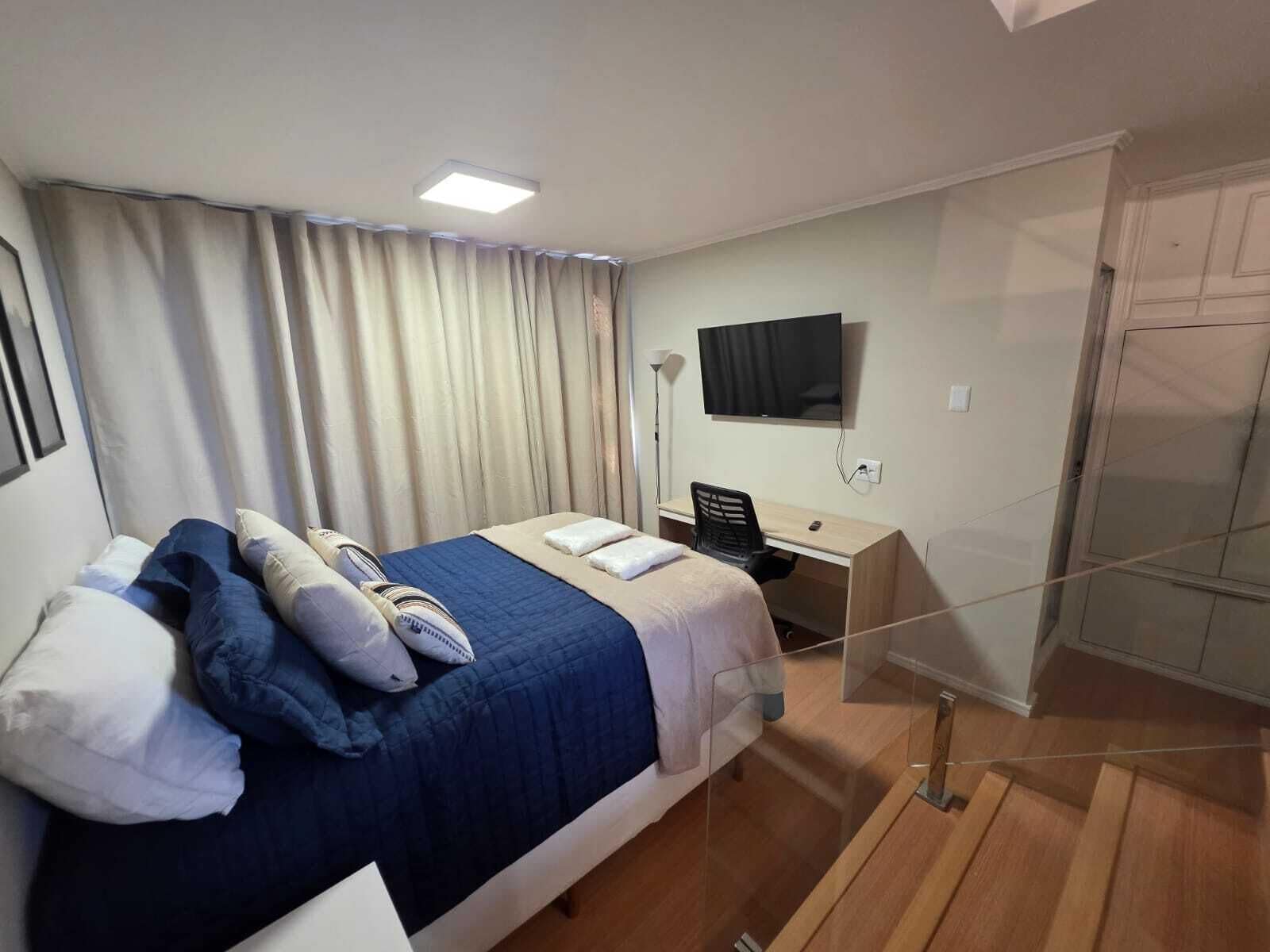 Quarto de hotel com cama, escrivaninha, cadeira, televisão e guarda-roupa.
