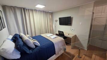 Quarto de hotel com cama, escrivaninha, cadeira, televisão e guarda-roupa.