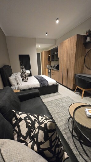 A modern bedroom with a bed, a sofa, a coffee table, and a kitchen area with wooden cabinets. -  Luxurious and Modern, Menlyn Maine Residences 825, Pretoria (Pretoria)