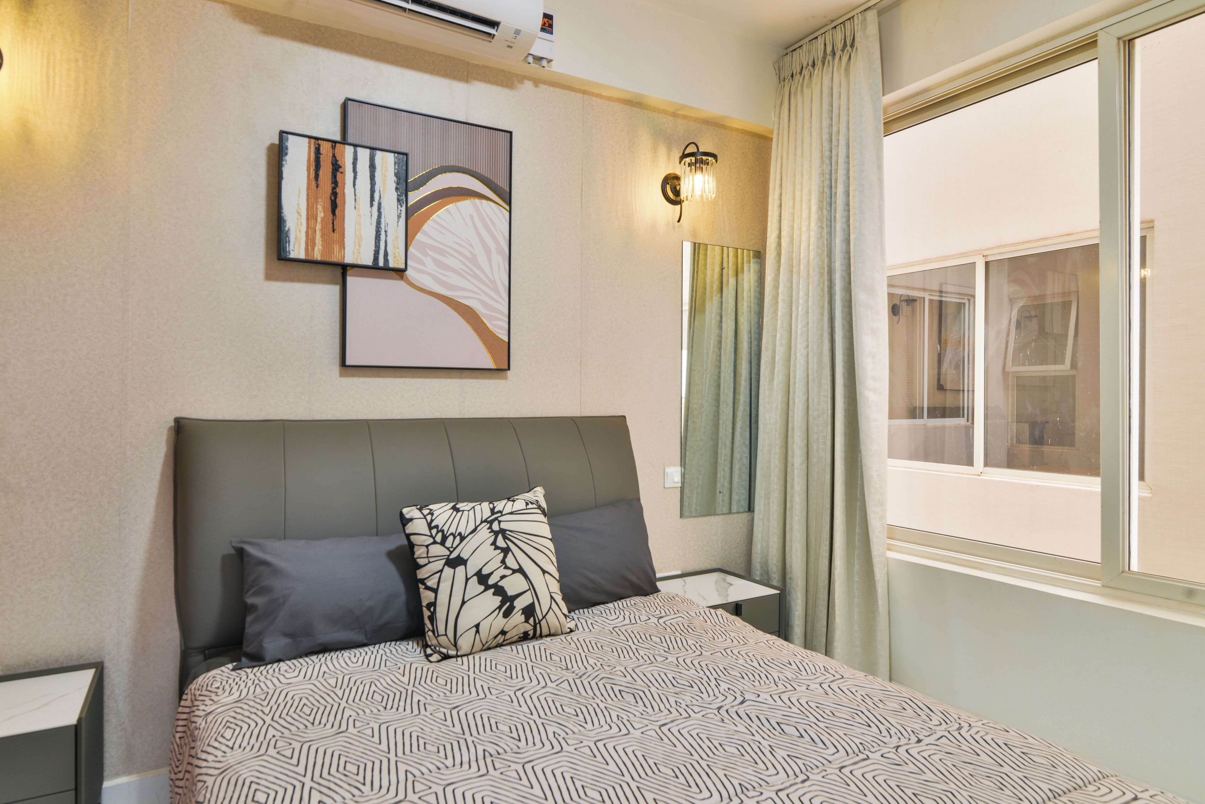 A hotel room with a bed, two pillows, a nightstand, a wall-mounted lamp, a window with curtains, and artwork on the wall.