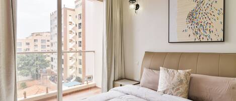 A hotel room with a large bed, a view of buildings through a window, and a wall-mounted light fixture.