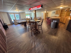 A spacious living area with a wooden dining table and chairs, a sofa, and a kitchen area with wooden cabinets and a refrigerator.