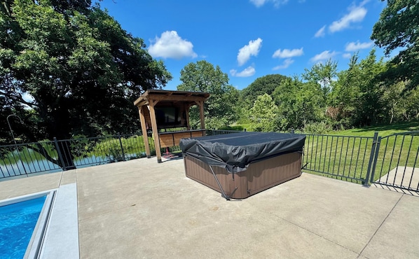 A backyard with a hot tub, a wooden pergola, a swimming pool, and a fenced area with trees.