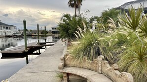 Una passerella in cemento con una panchina in pietra, un porto turistico con barche e una fila di case sullo sfondo.