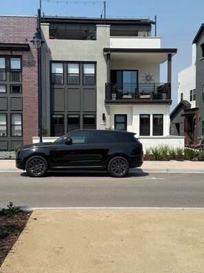 Un moderno edificio de dos plantas con balcón, amplias ventanas y un SUV negro estacionado adelante.