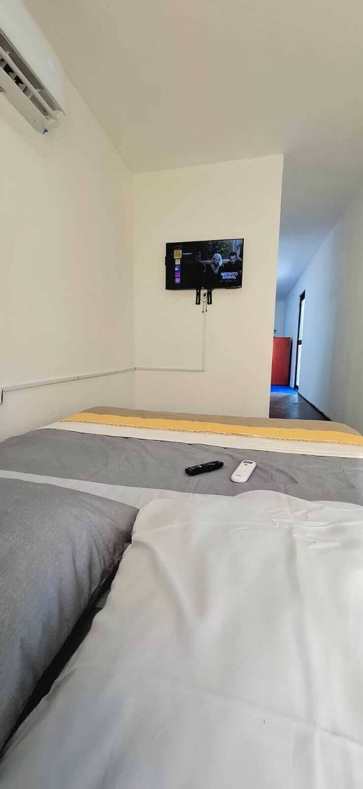 Una habitación con una cama, un televisor montado en la pared y un control remoto sobre la cama.