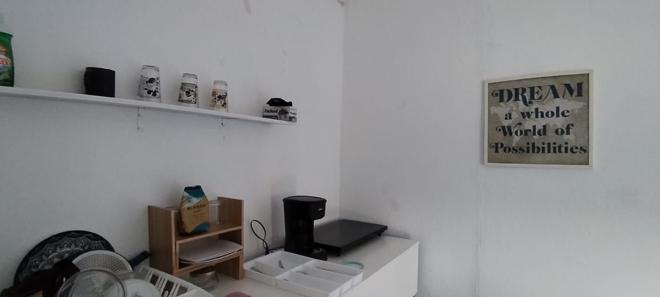 Una habitación con pared blanca, estante con varios objetos, cafetera y un cartel motivacional.