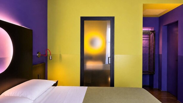 A hotel room with a bed, a door with a bright light, and a hallway with purple walls.