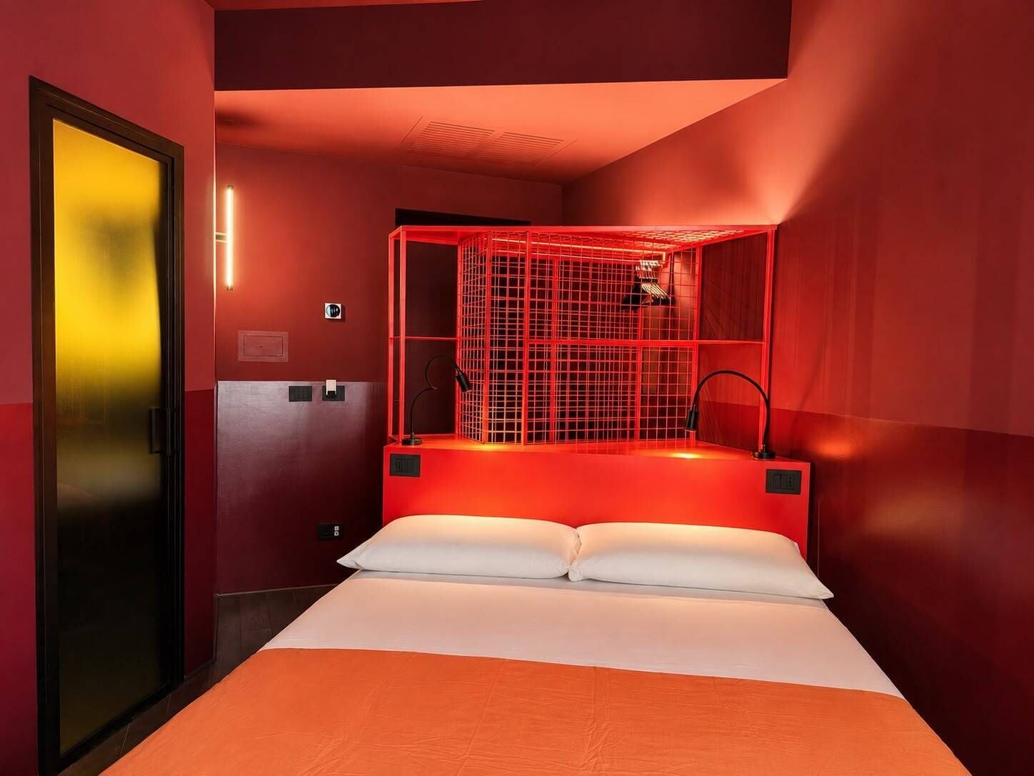 Une chambre d’hôtel avec un lit, un coin salon éclairé de lumière rouge et une porte dotée d’un panneau en verre dépoli.