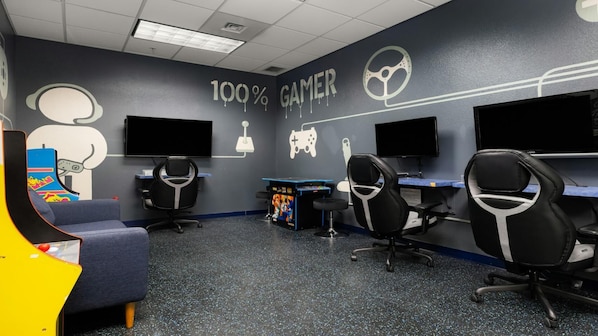 Ein Gaming-Raum mit vier Gaming-Stationen, jeweils ausgestattet mit einem Bildschirm und einem Stuhl. Die Wände sind dunkel gestaltet und zeigen Gaming-Grafiken sowie den Text "100% GAMER".