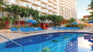 Ein Hotelpoolbereich mit blauen Liegestühlen und Sonnenschirmen, umgeben von Palmen und einem mehrstöckigen Gebäude.