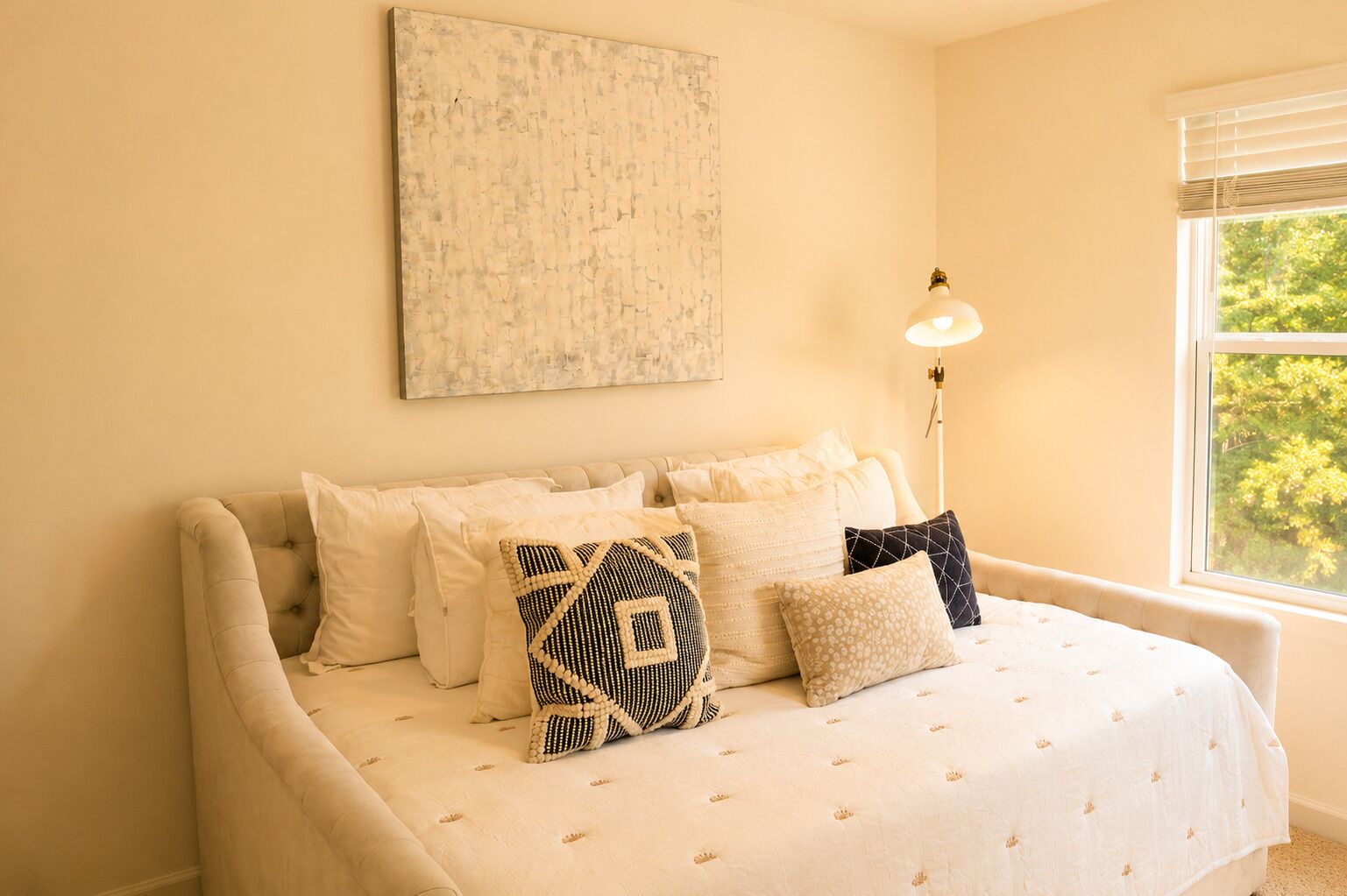 A bedroom with a bed, multiple pillows, a bedside lamp, a window with a view, and a piece of wall art.